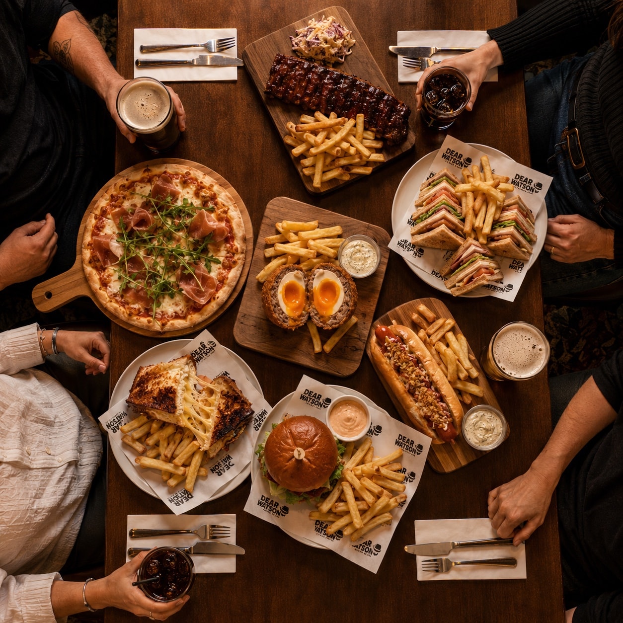 Overhead Dear Watson table spread with ribs, pinsa, sandwich, burger, hot dog, fries, and drinks.